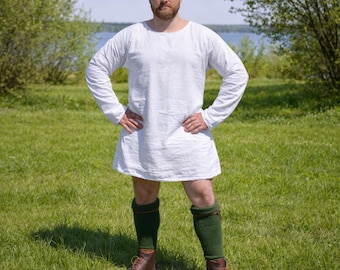 May include: A white long-sleeved tunic with a round neckline. The tunic is paired with green knee-high socks and brown leather boots. The person is standing on a grassy field.