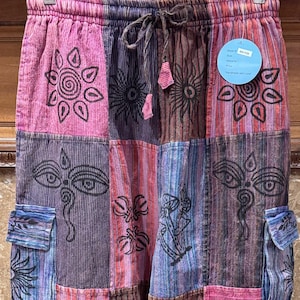 May include: Patchwork cargo pants featuring a drawstring waist and a multi-colored striped pattern in shades of pink, purple, blue, and brown. The fabric is printed with black line art designs including lotus flowers, eyes, and mushrooms.