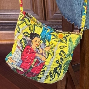 May include: A yellow crossbody bag with a colorful strap, featuring a vibrant print of a woman in a red dress. The bag is made of quilted fabric and is hanging on a blue chair.