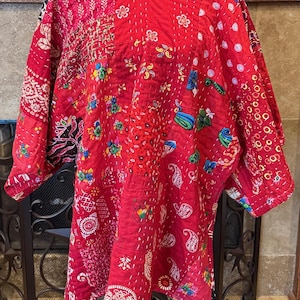 May include: A red patchwork tunic featuring a variety of colorful floral, paisley, and geometric patterns. The garment is constructed with visible white running stitch quilting details throughout, a round neckline, and wide, loose-fitting sleeves.