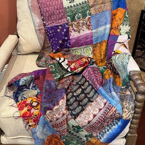 May include: A patchwork quilt draped over a beige armchair. The quilt features a variety of colorful fabric squares in shades of blue, purple, orange, and green. The quilt's design includes floral and geometric patterns, creating a vibrant and textured appearance.