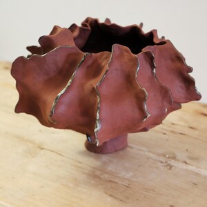 Peut inclure: Un vase en céramique de couleur terre cuite fait à la main, avec un design unique et froissé. Le vase a un large sommet ouvert et une base en piédestal. Les bords des volants sont accentués d'une glaçure dorée métallique.