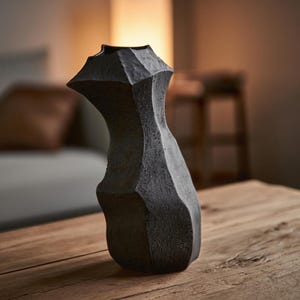 Peut inclure: Un vase texturé gris foncé avec un design angulaire et géométrique. Le vase a une forme asymétrique unique et repose sur une surface en bois. L'intérieur n'est pas visible. L'arrière-plan est flou, suggérant un cadre de salon.