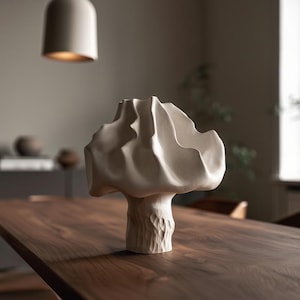 Peut inclure: Un vase en céramique abstrait de couleur crème avec un design organique texturé. Le vase a un dessus large et froissé et une base texturée en forme de tronc d'arbre. Il repose sur une table en bois foncé, avec un fond aux tons neutres.