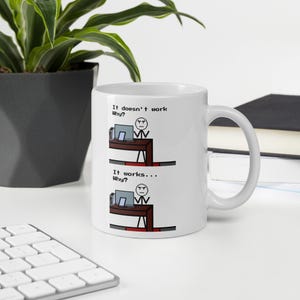 Op de afbeelding: Witte keramische mok met een humoristische computergerelateerde afbeelding. De mok toont een stokfiguur aan een bureau met de tekst "It doesn't work Why?" en "It works... Why?"