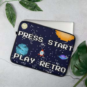 Può includere: Una custodia per laptop blu navy con un design pixelato a tema spaziale. La custodia presenta una luna, un pianeta, un razzo e le parole "PRESS START PLAY RETRO" in lettere bianche. La custodia è progettata per un laptop.