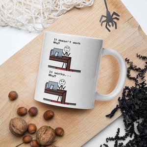 Può includere: Tazza in ceramica bianca con una grafica umoristica a tema informatico. Il design mostra una persona pixelata a una scrivania con il testo "It doesn't work Why?" e "It works... Why?" La tazza ha un manico bianco.