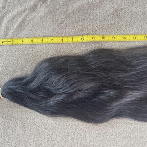 Puede incluir: Un mech&oacute;n de cabello oscuro, asegurado con una goma el&aacute;stica, descansa sobre una superficie beige. Una cinta m&eacute;trica amarilla se coloca a lo largo del cabello, midiendo unos 46 cm.