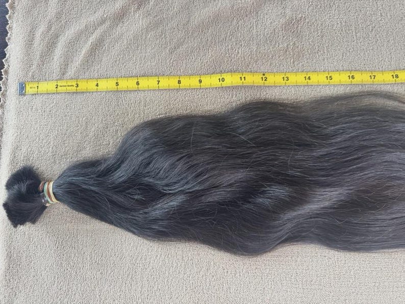 Puede incluir: Un mech&oacute;n de cabello oscuro, asegurado con una goma el&aacute;stica, descansa sobre una superficie beige. Una cinta m&eacute;trica amarilla se coloca a lo largo del cabello, midiendo unos 46 cm.