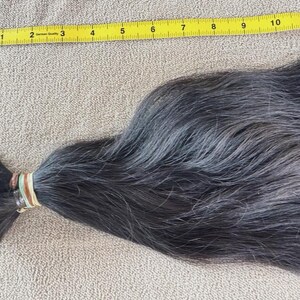 Puede incluir: Un mech&oacute;n de cabello gris oscuro, sujeto con una goma el&aacute;stica, junto a una cinta m&eacute;trica amarilla. El cabello mide aproximadamente 30 cm de largo, con una textura natural y ligeramente ondulada.