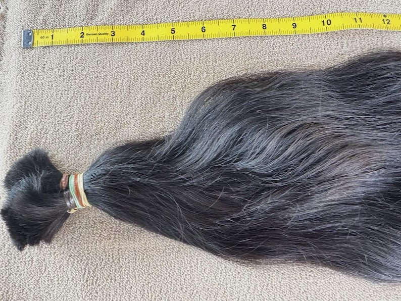 Puede incluir: Un mech&oacute;n de cabello gris oscuro, sujeto con una goma el&aacute;stica, junto a una cinta m&eacute;trica amarilla. El cabello mide aproximadamente 30 cm de largo, con una textura natural y ligeramente ondulada.