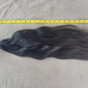 Puede incluir: Un mech&oacute;n de cabello negro oscuro, asegurado con una goma el&aacute;stica, descansa sobre una superficie de color neutro. Una cinta m&eacute;trica amarilla se coloca junto al cabello, midiendo unos 58 cm de largo.