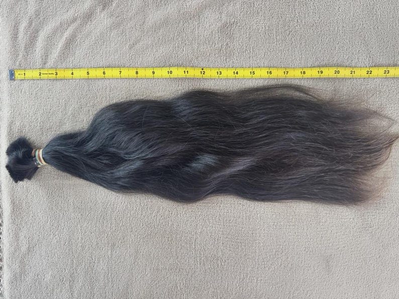Puede incluir: Un mech&oacute;n de cabello negro oscuro, asegurado con una goma el&aacute;stica, descansa sobre una superficie de color neutro. Una cinta m&eacute;trica amarilla se coloca junto al cabello, midiendo unos 58 cm de largo.
