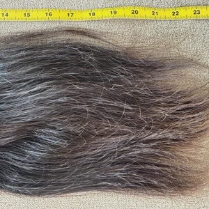 Puede incluir: Una secci&oacute;n de cabello gris oscuro y marr&oacute;n, con mechones plateados, se coloca junto a una cinta m&eacute;trica amarilla. El cabello mide aproximadamente 30 cm de largo, con una textura natural y variaci&oacute;n de color. El cabello es probablemente para una peluca o extensi&oacute;n.
