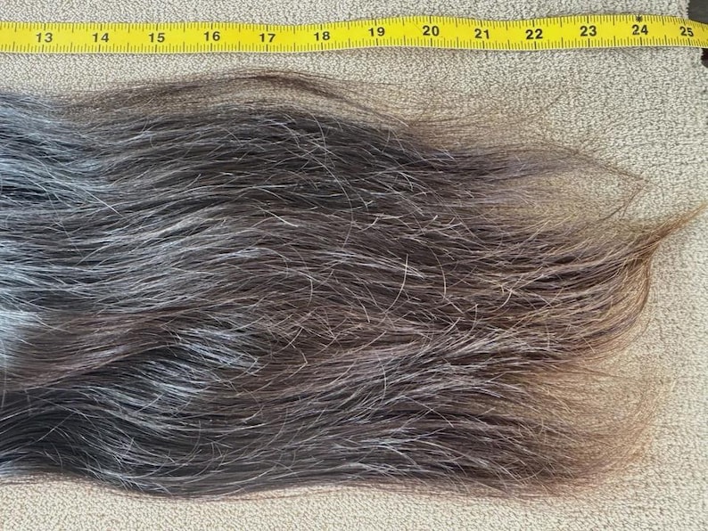 Puede incluir: Una secci&oacute;n de cabello gris oscuro y marr&oacute;n, con mechones plateados, se coloca junto a una cinta m&eacute;trica amarilla. El cabello mide aproximadamente 30 cm de largo, con una textura natural y variaci&oacute;n de color. El cabello es probablemente para una peluca o extensi&oacute;n.
