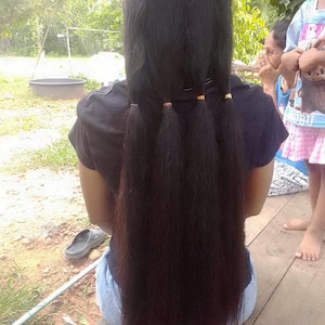 Puede incluir: Cabello largo y oscuro peinado con tres peque&ntilde;as coletas sujetas con gomas para el pelo naranjas y doradas. El cabello cae por la espalda de una persona que lleva una camiseta negra y vaqueros azules. El fondo incluye una superficie de madera y vegetaci&oacute;n.