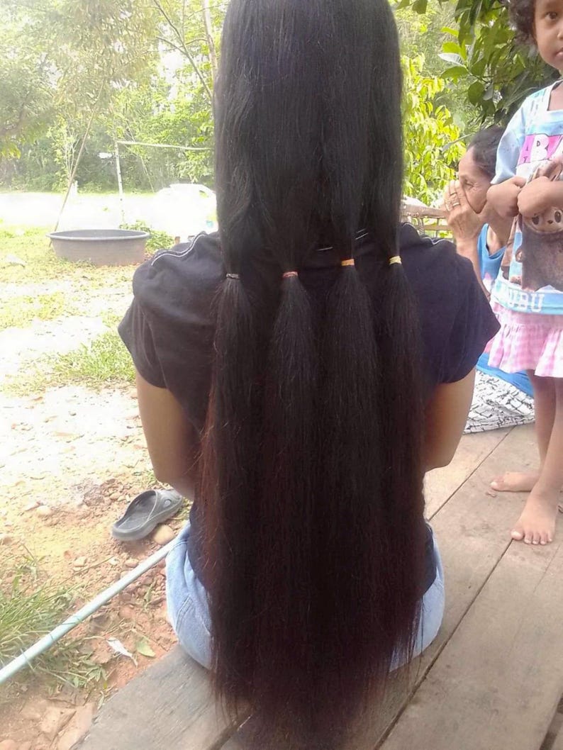 Puede incluir: Cabello largo y oscuro peinado con tres peque&ntilde;as coletas sujetas con gomas para el pelo naranjas y doradas. El cabello cae por la espalda de una persona que lleva una camiseta negra y vaqueros azules. El fondo incluye una superficie de madera y vegetaci&oacute;n.