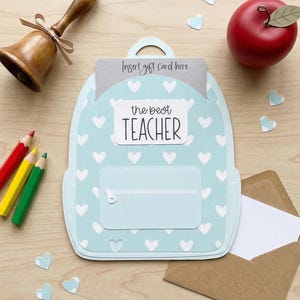 May include: A light blue backpack-shaped gift card holder with white heart patterns and the text "the best TEACHER." Includes a red apple, pencils, a bell, and a brown envelope with a white card.