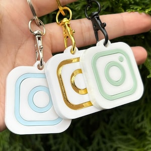 May include: Three white square keyrings featuring Instagram logo designs. Each keyring has a different colour accent: light blue, gold, and green. The keyrings have metal clasps and key rings.