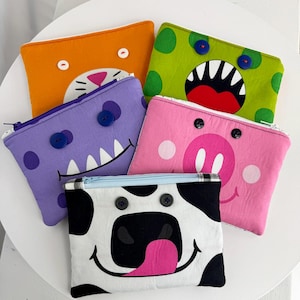 May include: Five colorful, zippered pouches with whimsical animal faces. The pouches feature an orange cat, green monster, purple monster, pink pig, and a black and white cow. Each pouch has button eyes and a unique design.