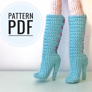 Może przedstawiać: Szydełkowe buty dla lalek w kolorze jasnoniebieskim z różowymi akcentami. Buty na wysokim obcasie mają sznurowanie z tyłu. Tekst "PATTERN PDF" znajduje się w niebieskim okręgu. Nogi lalki są różowe.