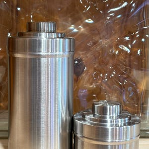 May include: Two stainless steel containers with a brushed metal finish. One is tall and cylindrical, the other is shorter and wider. Both have a stepped, ridged top. The containers are likely for storage.