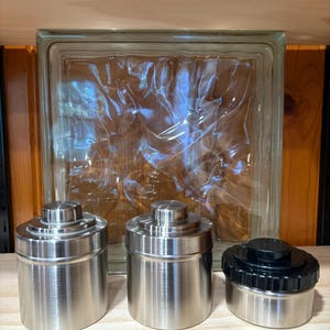 May include: Three stainless steel canisters with lids are displayed on a wooden shelf. Two canisters have matching silver lids, while the third has a black lid. A clear glass block is in the background.