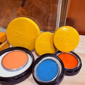 May include: A collection of vintage Kodak camera filters and lens caps. The yellow caps are embossed with the Kodak logo. The filters are in black holders with orange and blue colored glass. The items are arranged on a wooden surface.