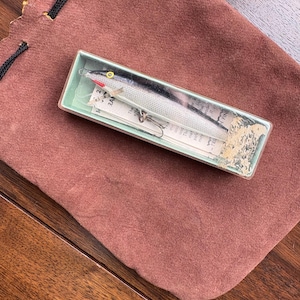 May include: A vintage fishing lure in a clear plastic box, resting on a brown suede pouch. The lure is silver and black with a red accent. The box has a label with text. The pouch is closed with a black drawstring.