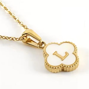 Könnte beinhalten: Goldfarbene Halskette mit einem kleeblattförmigen Anhänger. Der Anhänger hat eine weiße Mitte und den Buchstaben "L". Der Anhänger ist goldfarben umrandet und hängt an einer zarten Goldkette. Die Halskette ist vor weißem Hintergrund.