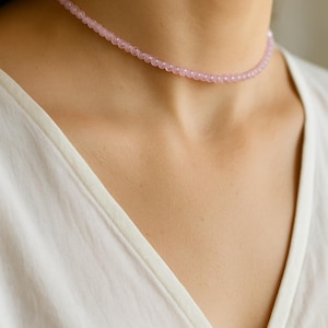May include: A delicate pink beaded choker necklace. The necklace is composed of small, round, light pink beads strung together to form a close-fitting necklace. The necklace is worn against a white shirt.