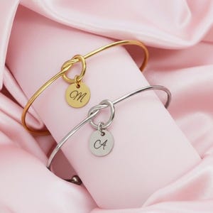 May include: Two elegant bangles, one gold and one silver, are displayed on a pink surface. Each bangle has a knot design and a small, circular charm with an initial engraved. The gold bangle has the letter "M", and the silver one has "CA".