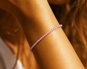 May include: A delicate, light pink beaded bracelet. The bracelet is composed of small, round beads strung together. The jewelry is worn on a tanned arm, with the focus on the bracelet's subtle color and design. The bracelet is a simple, elegant accessory.