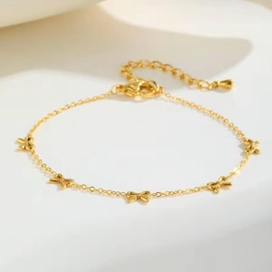 May include: A delicate gold-colored bracelet featuring small bow charms. The adjustable chain bracelet is made of a shiny, reflective metal. The bracelet is a jewelry item.