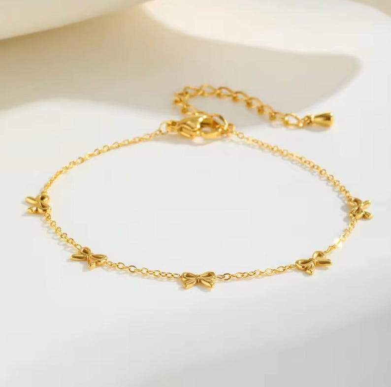 May include: A delicate gold-colored bracelet featuring small bow charms. The adjustable chain bracelet is made of a shiny, reflective metal. The bracelet is a jewelry item.