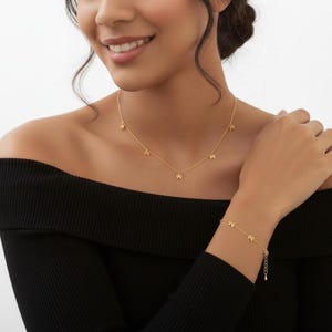 May include: A gold-colored necklace and bracelet set. The necklace features five small star charms, and the bracelet has a similar design. The jewelry is worn against a black off-the-shoulder top.
