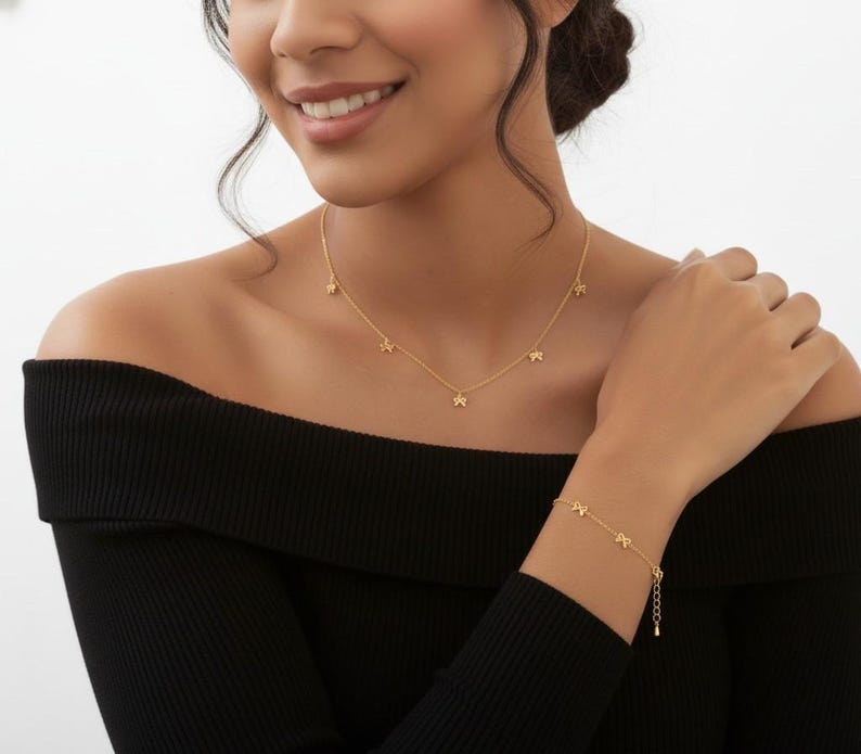 May include: A gold-colored necklace and bracelet set. The necklace features five small star charms, and the bracelet has a similar design. The jewelry is worn against a black off-the-shoulder top.