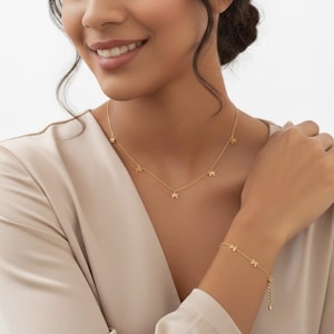 May include: A gold-colored necklace and bracelet set. The necklace features small star-shaped charms evenly spaced along the chain. The matching bracelet has similar star charms. The jewelry is worn against a neutral-colored top.