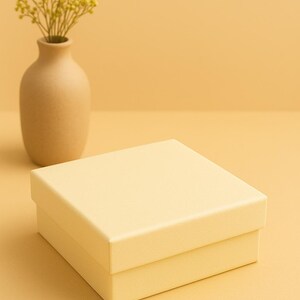 May include: A square, pale yellow gift box with a matching lid sits on a pale yellow surface. A small, tan vase holds a bouquet of yellow flowers, set against a pale yellow background.
