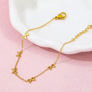 May include: Gold-colored bracelet featuring a delicate chain with bow-shaped charms. The bracelet has a lobster clasp closure and is displayed on a pink surface with a white dish in the background. The bow charms are evenly spaced along the chain.