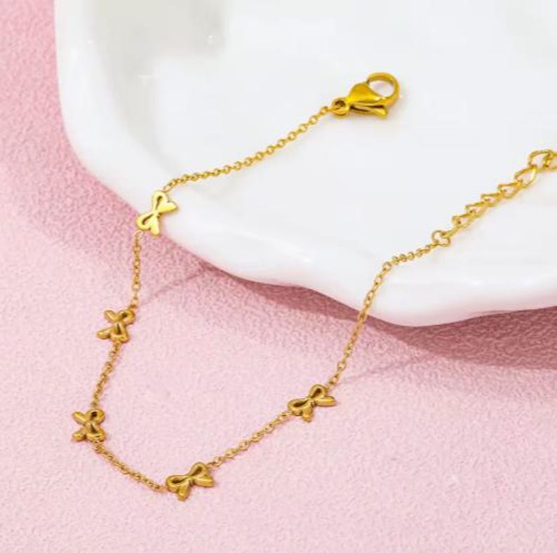 May include: Gold-colored bracelet featuring a delicate chain with bow-shaped charms. The bracelet has a lobster clasp closure and is displayed on a pink surface with a white dish in the background. The bow charms are evenly spaced along the chain.