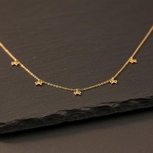 May include: A delicate gold-colored necklace featuring five small bow-shaped charms. The necklace is displayed against a dark background, highlighting the fine details of the jewelry. The chain is thin and elegant, suitable for everyday wear.