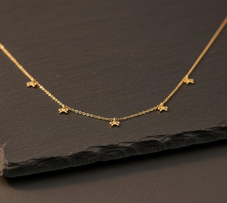 May include: A delicate gold-colored necklace featuring five small bow-shaped charms. The necklace is displayed against a dark background, highlighting the fine details of the jewelry. The chain is thin and elegant, suitable for everyday wear.