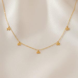 May include: Delicate gold-colored necklace featuring small bow-shaped charms evenly spaced along the chain. The necklace is displayed on a soft, white fabric background, highlighting the jewelry's elegant design. A simple and stylish accessory.