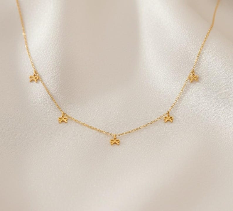 May include: Delicate gold-colored necklace featuring small bow-shaped charms evenly spaced along the chain. The necklace is displayed on a soft, white fabric background, highlighting the jewelry's elegant design. A simple and stylish accessory.
