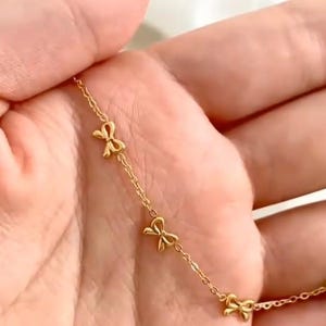 May include: Delicate gold-colored necklace featuring a series of small bow-shaped charms. The necklace is displayed against a soft, neutral background, highlighting the jewelry's intricate design and warm hue. The chain is thin and elegant.