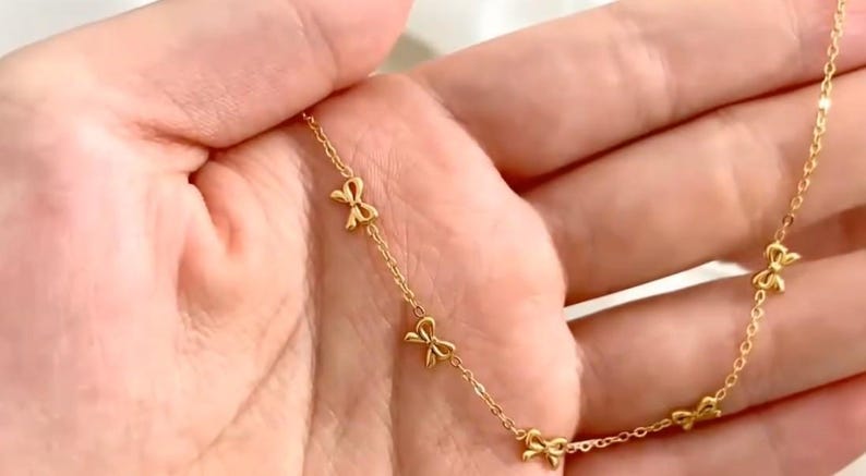May include: Delicate gold-colored necklace featuring a series of small bow-shaped charms. The necklace is displayed against a soft, neutral background, highlighting the jewelry's intricate design and warm hue. The chain is thin and elegant.