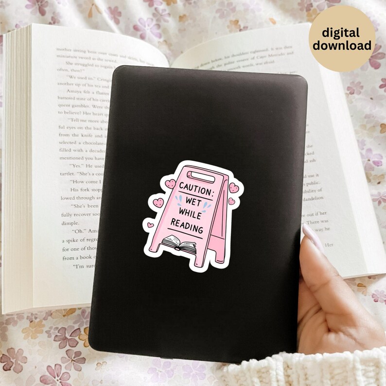 May include: A pink sticker on a black surface with the text "CAUTION: WET WHILE READING" in white. The sticker is shaped like a caution sign with hearts and a book illustration. The words "digital download" are in the upper right corner.