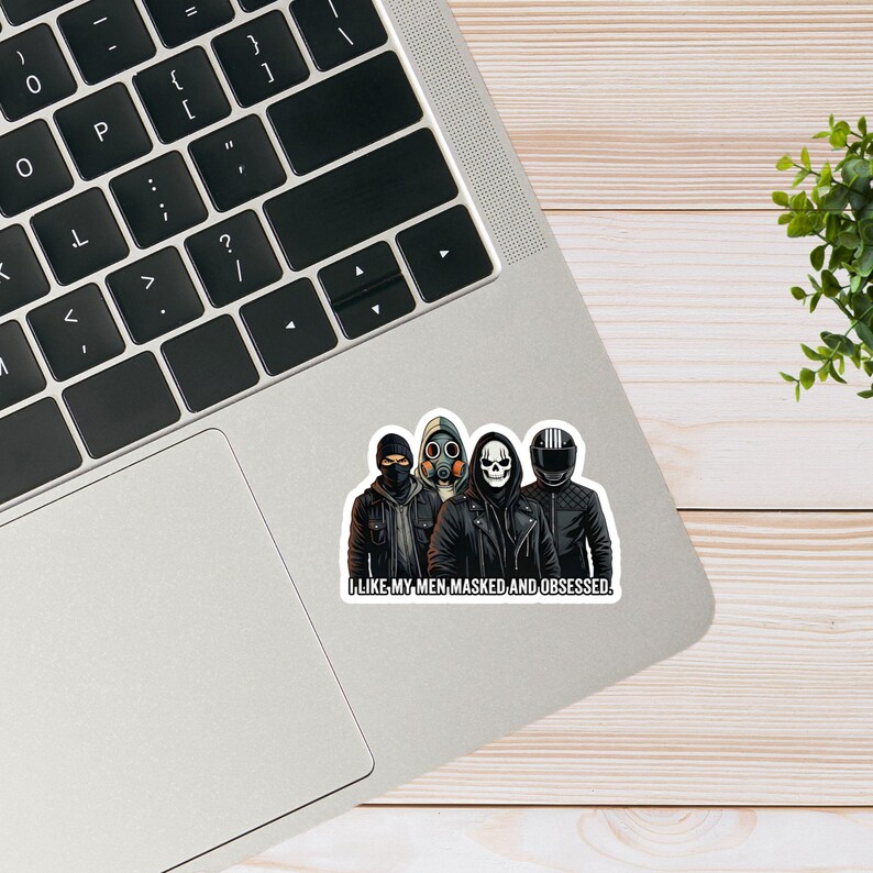 May include: A sticker featuring four figures in black jackets and masks, including a skull, gas mask, and motorcycle helmet. The text "I LIKE MY MEN, MASKED, AND OBSESSED" is at the bottom. The sticker is on a laptop.