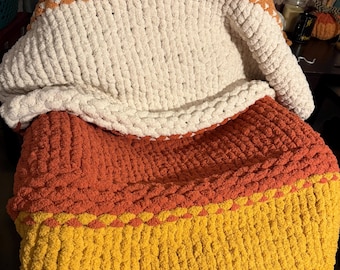 Puede incluir: Una manta de punto grueso en los colores del maíz dulce: naranja, crema y amarillo. La manta está doblada, mostrando las diferentes secciones de color. La textura del hilo es visible, creando un aspecto acogedor y acogedor.